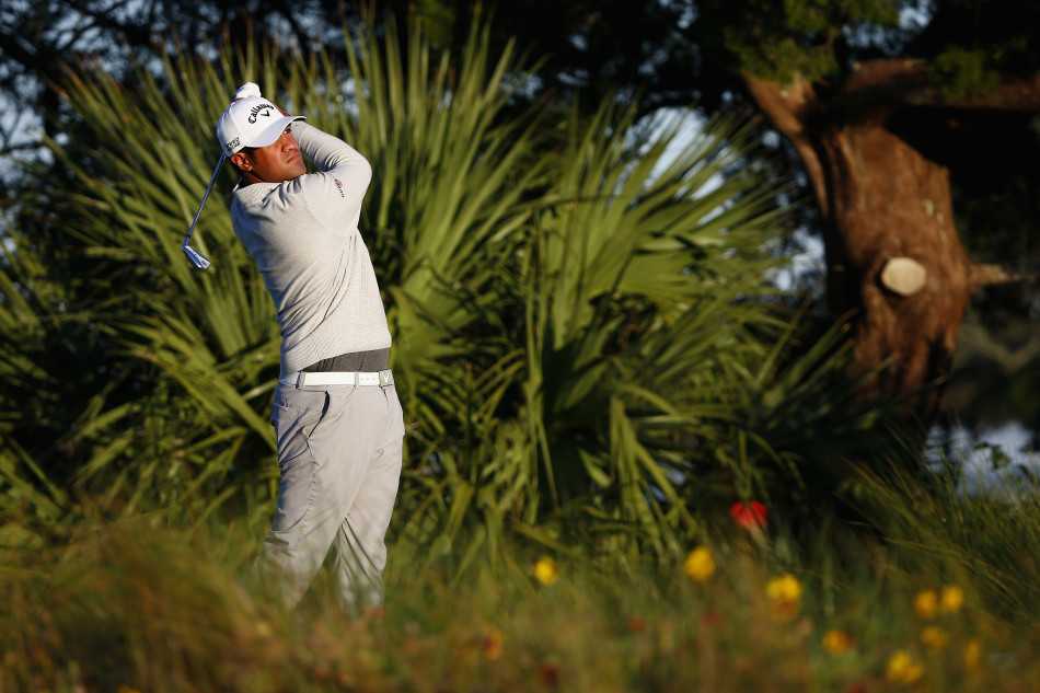 球员在沙坑中救球后，沙子在空中划出一道金色的弧线，与草地的翠绿和天空的蔚蓝构成一幅充满力量感的画面。