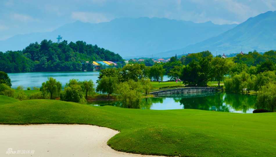 从高处俯瞰，一条优美的球道沿湖岸蜿蜒，湖水碧绿，倒映着天空，沙坑像白色贝壳点缀在绿地中。
