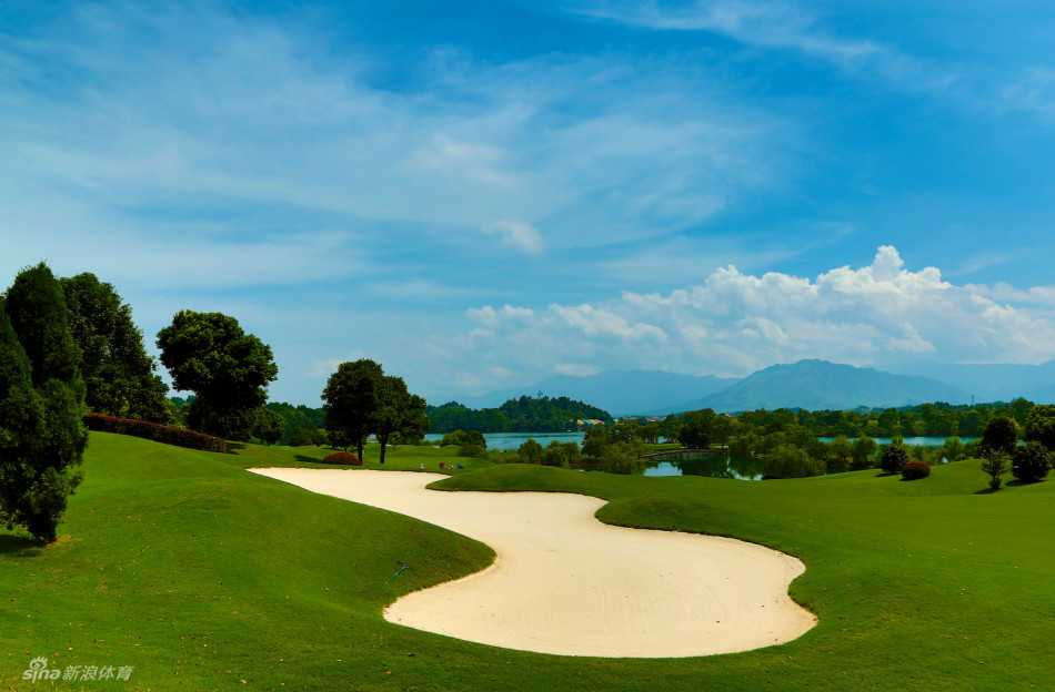 一位球手正在水障碍前的发球台凝神准备开球，白色的球车停在身后，对岸的球道绿意盎然。