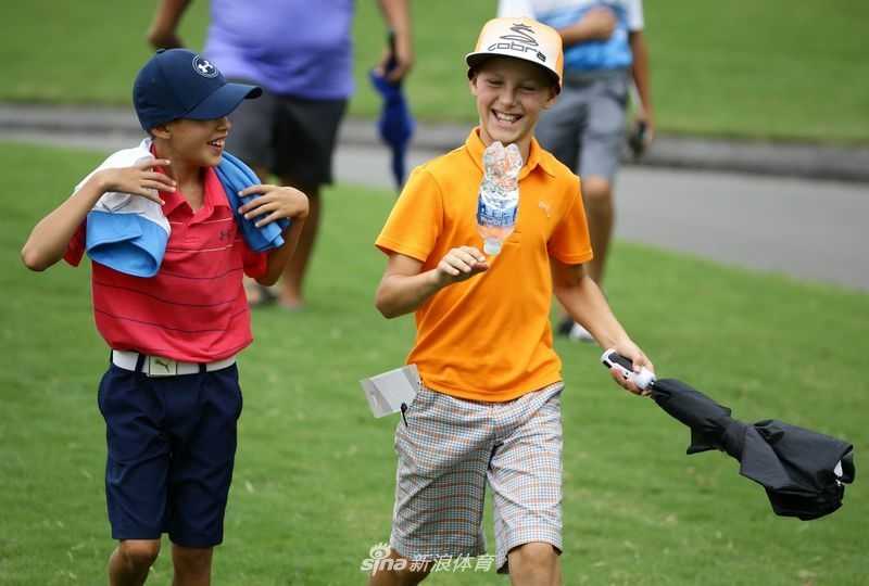 著名高尔夫球星瑞奇·福勒俯身围栏边，面带他标志性的友善微笑，正在一顶帽子上为小球迷签名。