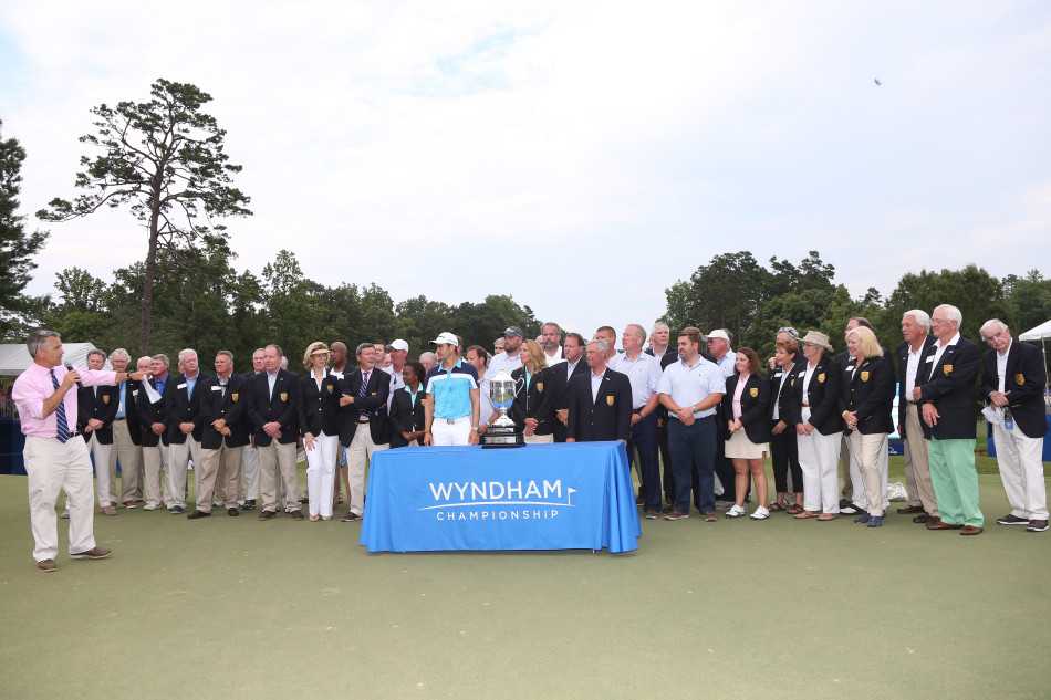 冠军毕耶加斯与赛事官员并肩站立，手持奖杯面向镜头微笑，身后绿茵赛场与蓝天构成完美胜利背景